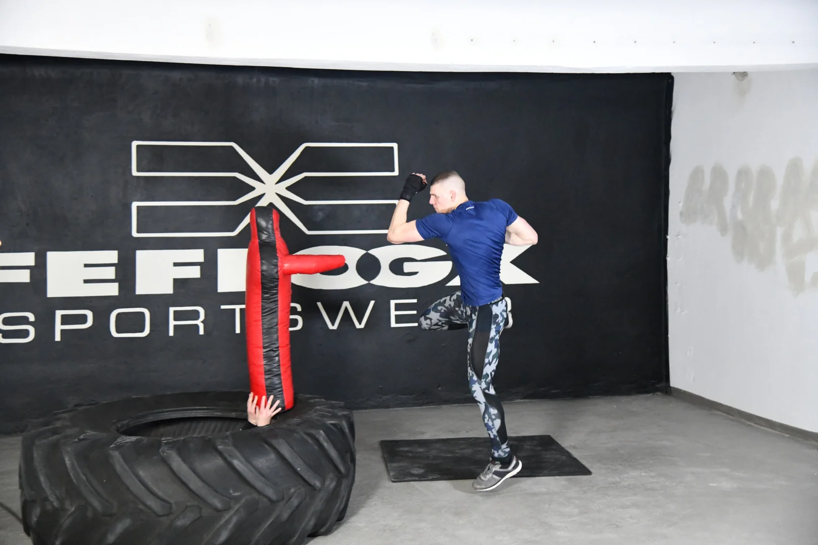 FEFLOGX, MMA-Training & Urban Functional Fitness Shooting in FFX Sport Leggings Camouflage, FFX Sport Shorts, FFX Boxbandagen & Sport Sweater, Fitness Longsleeve mit Kampfsport Dummy & Sport Reifen mit Hammer schlagen, Flying Superman Punch KO-Schlag Boxen in Industrie Ruine in Frankfurt am Main, mit FEFLOGX Logo-Graffiti an der Wand (3).